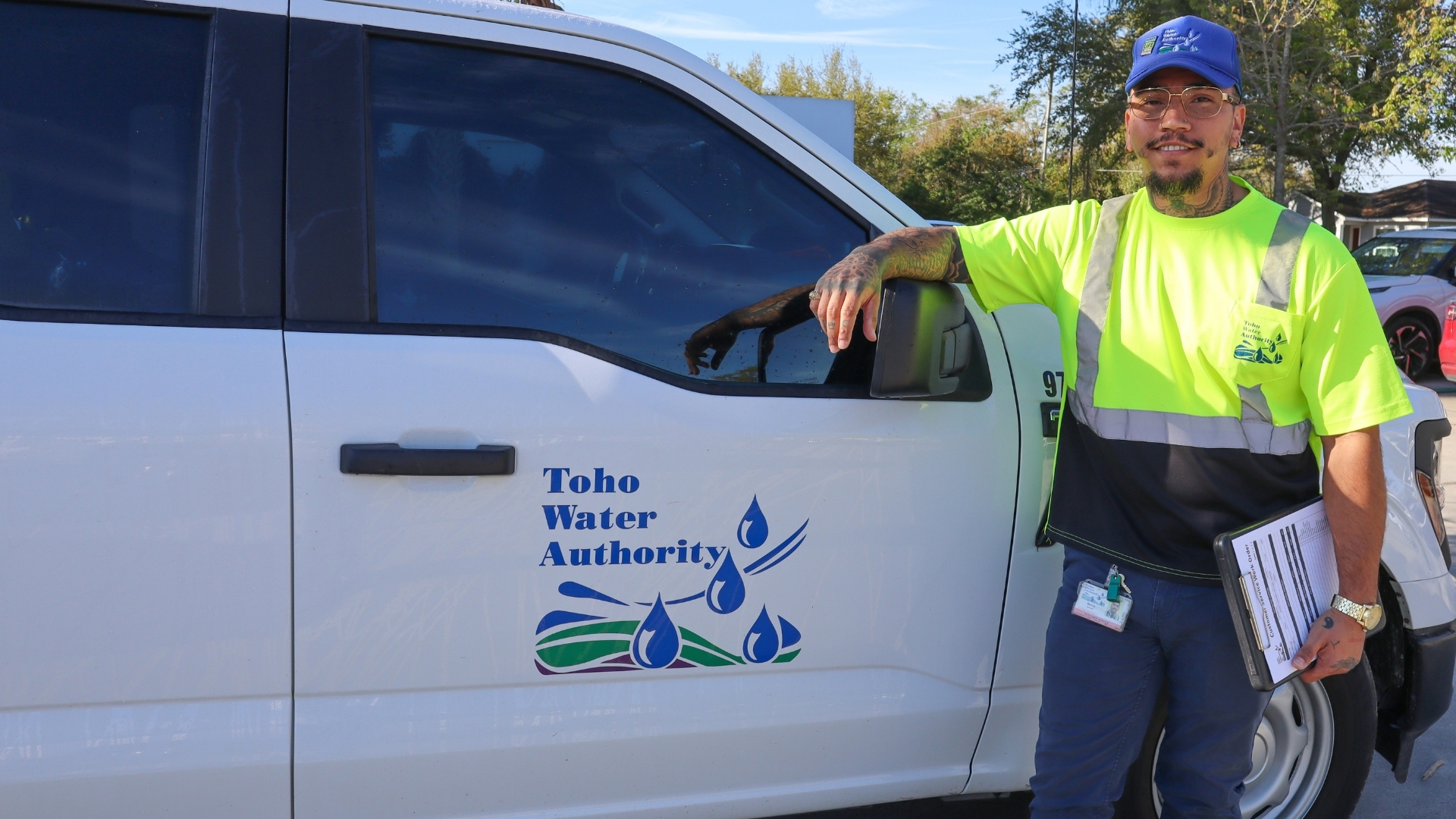 Toho employee wearing branded reflective attire, identification badge, holding a clipboard leans against a toho water authority branded truck