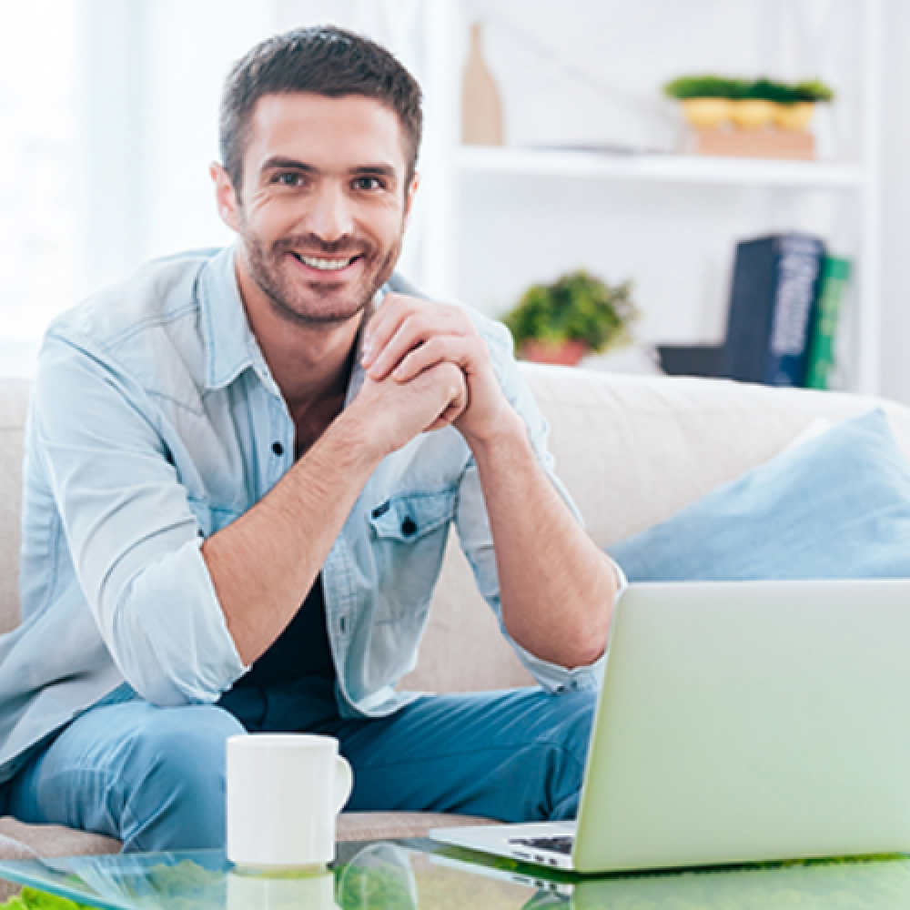 Image of a smiling man with a laptop sitting in a living room.