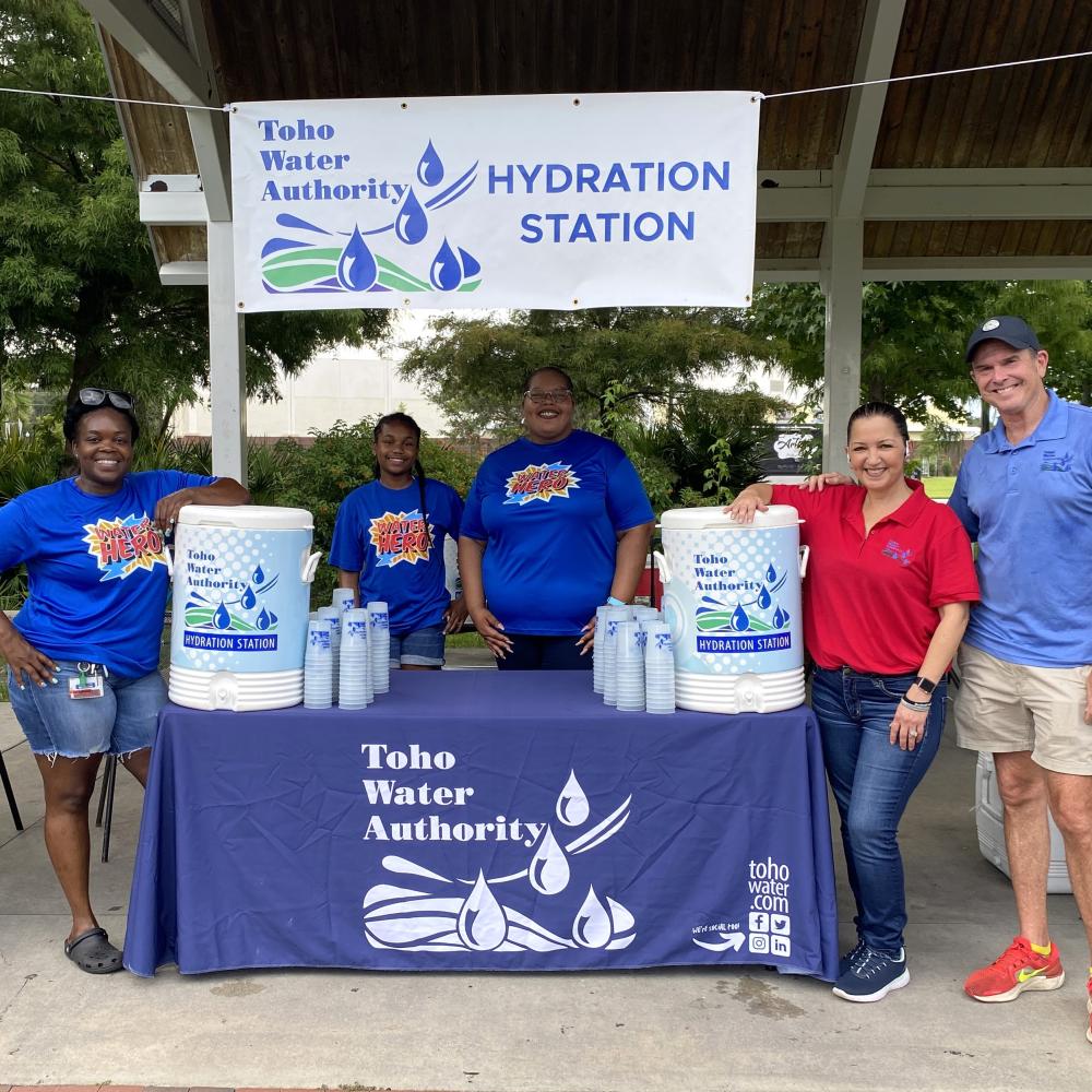 Photo of five individuals representing Toho Water an an event. Toho water coolers are in the photo along with plastic cups. A banner with the Toho logo and the words "Hydration Station" is hanging. 